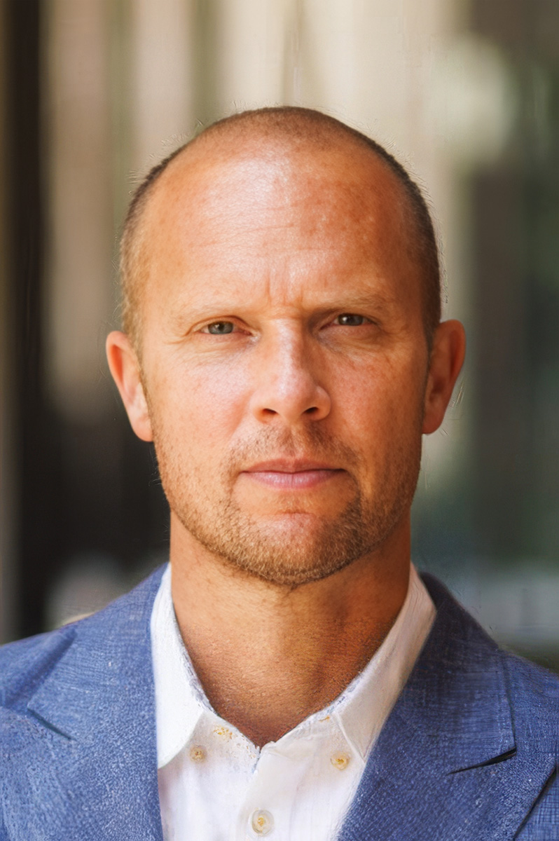 Norman Spaulding in a blue suit and white shirt stands against a blurred background.