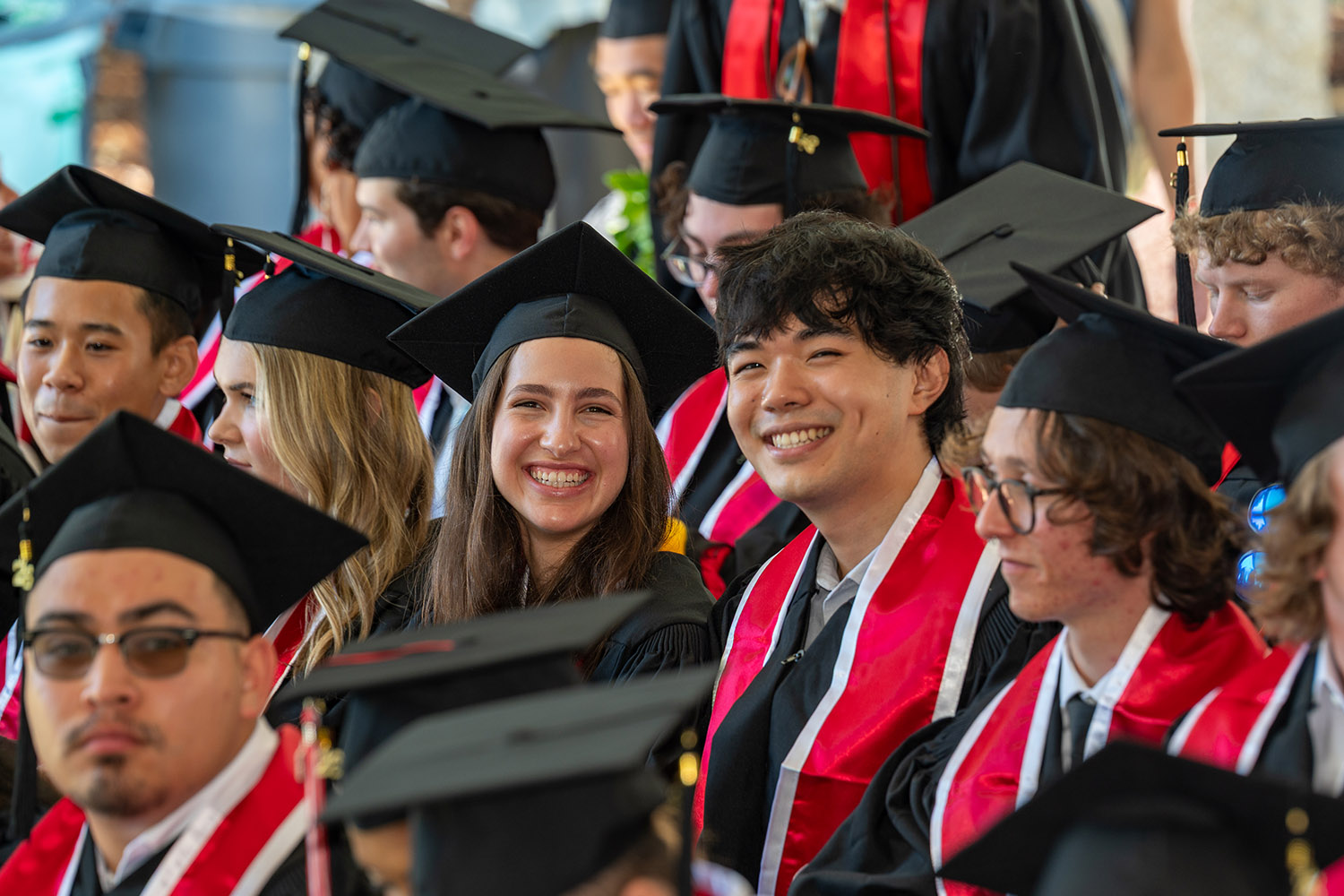 Graduates in caps and gowns are seated together, eagerly awaiting their ceremony at an outdoor graduation event.