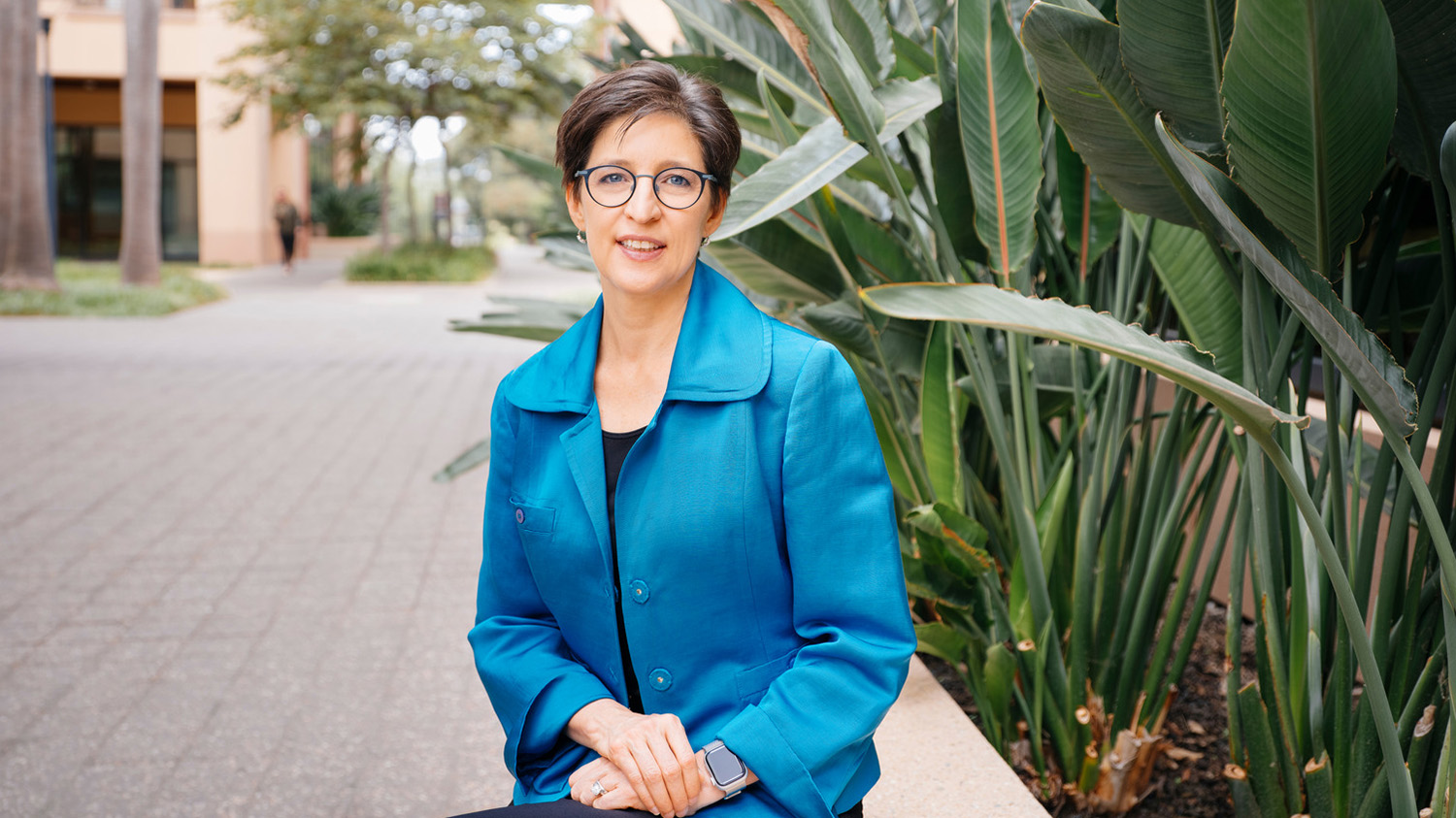 Susan Athey sitting on a bench near tropical plants outdoors on the Stanford campus.