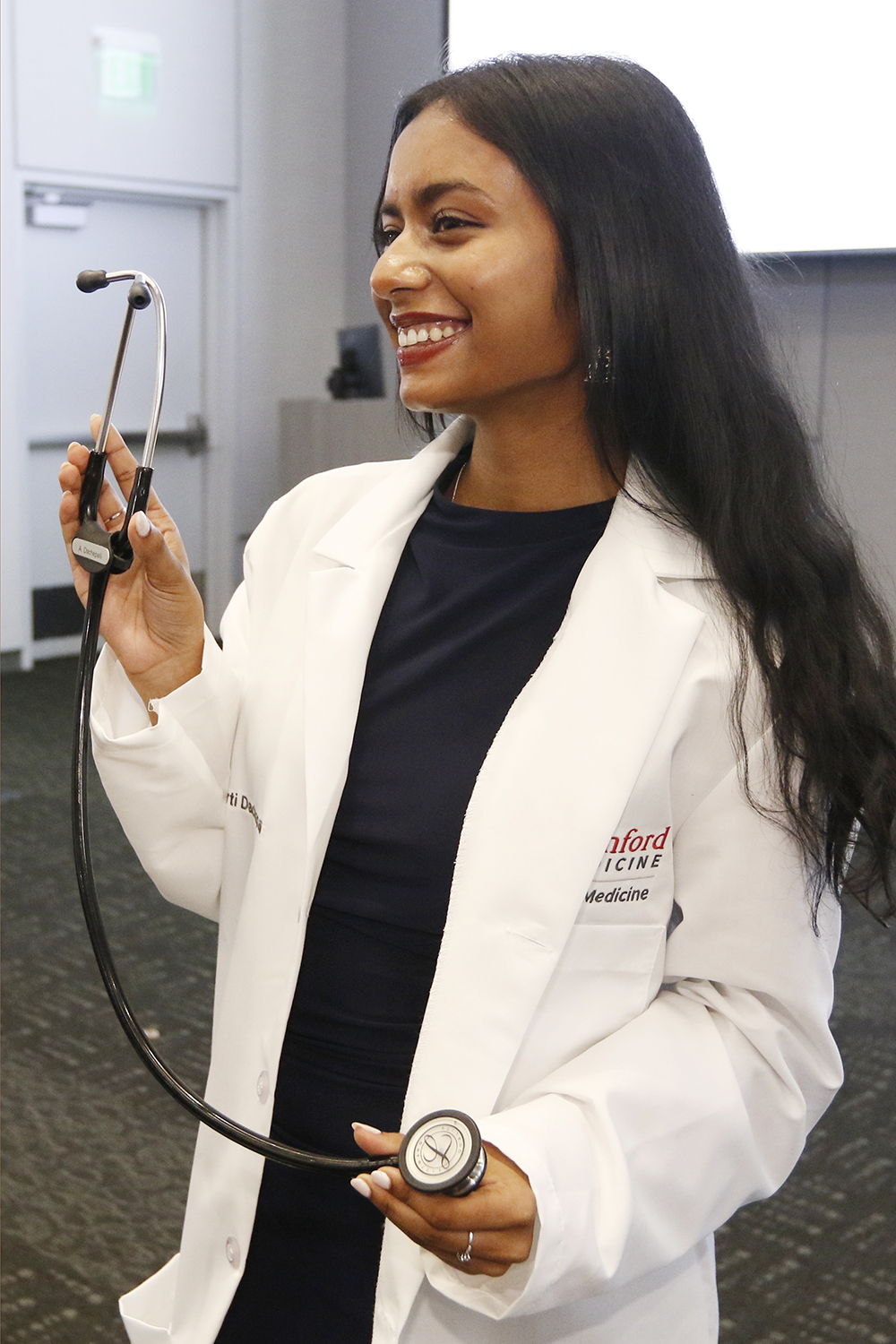 A medical student in a white coat holds up her stethoscope, smiling during a ceremony.