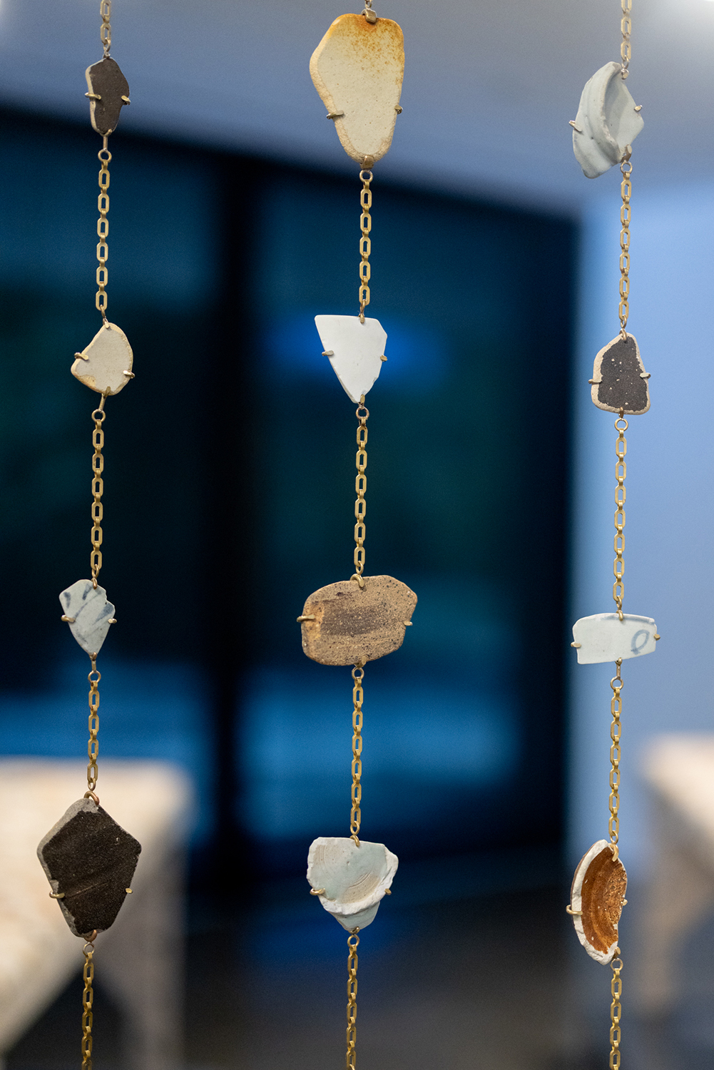 Gold chains hang vertically, each strung with fragments of ceramic and pottery held in small metal prongs.