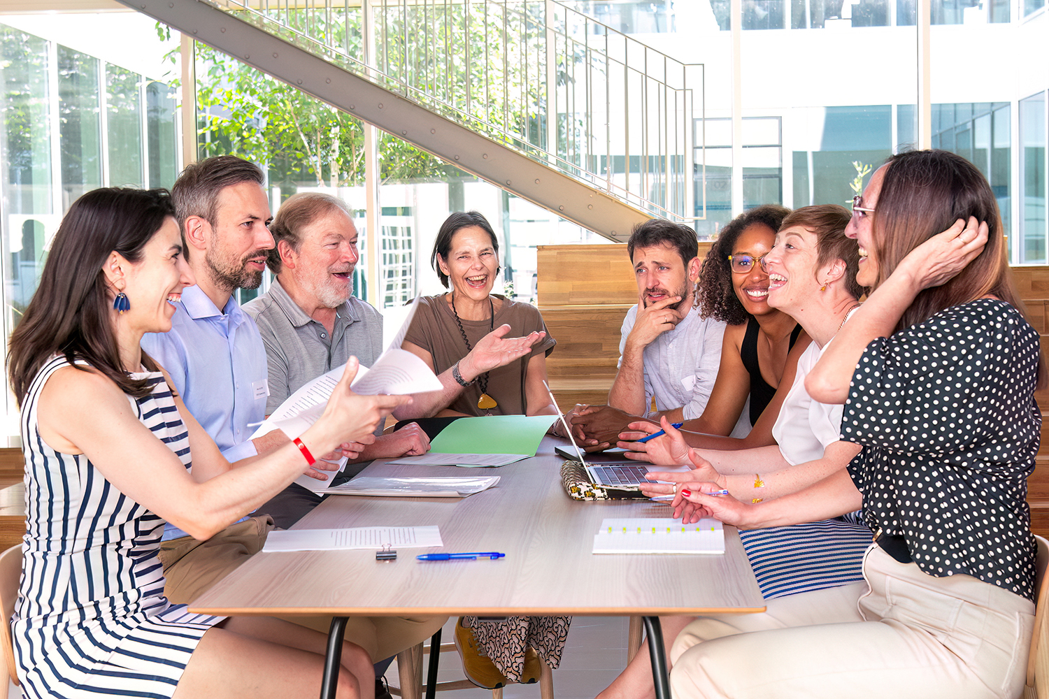 Eight colleagues laugh together around a table with papers and a laptop in a bright, glass-walled atrium.