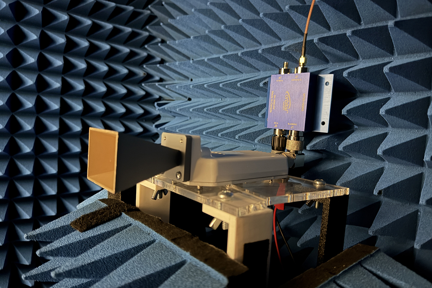 Horn antenna on a clear acrylic stand points toward a blue radio-frequency component mounted on the wall of a foam-lined chamber.