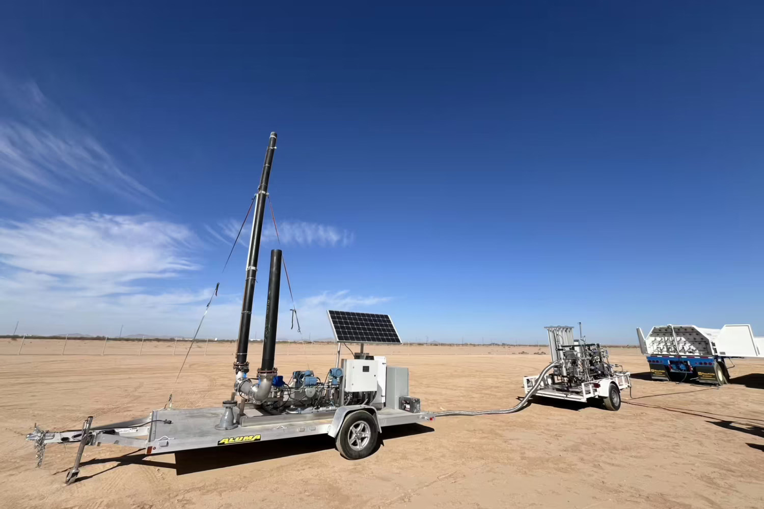 Trailer-mounted equipment with tall black pipes and a solar panel sits on open desert ground, hooked by a hose to a second trailer.
