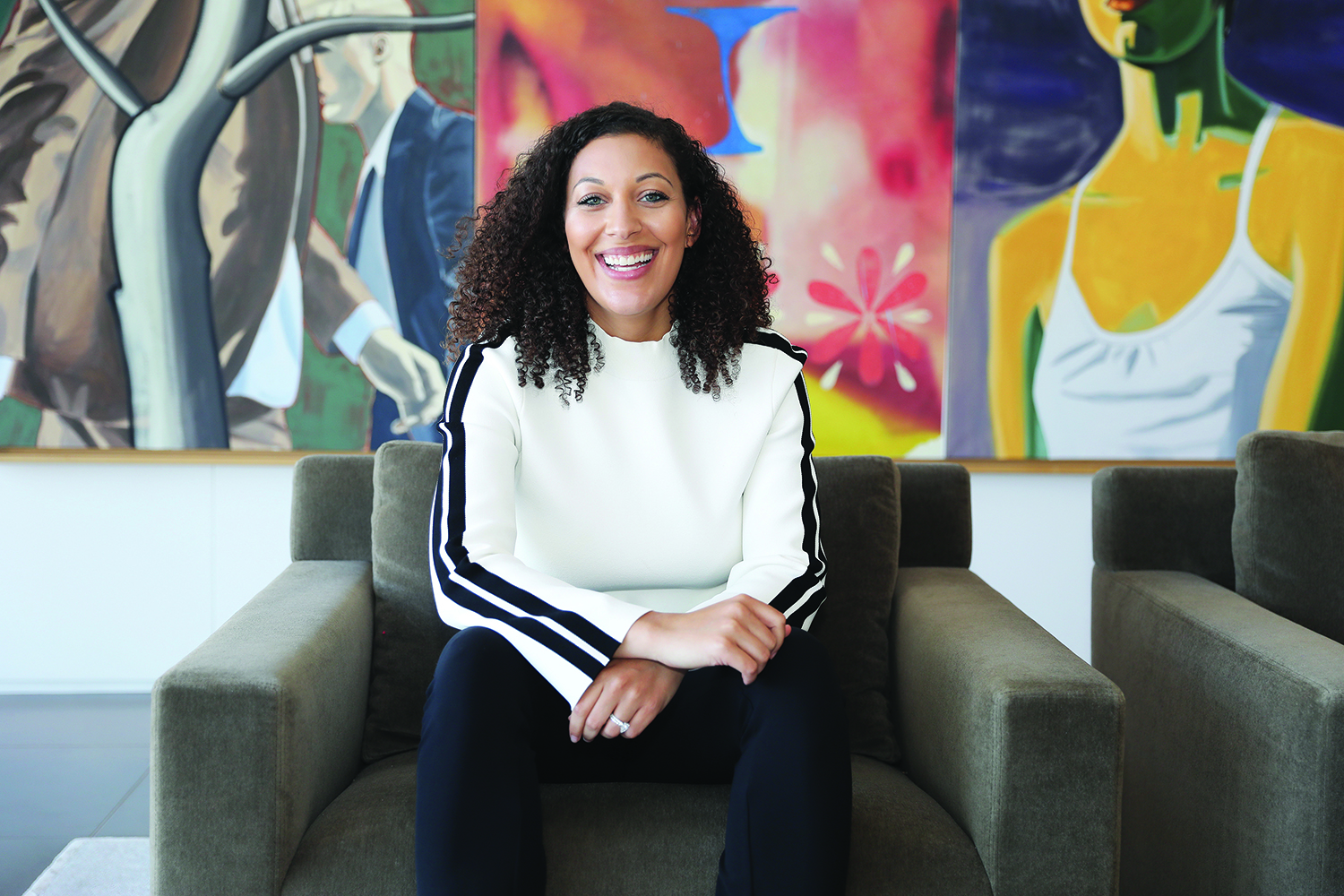 Krista Whitaker in a white sweatshirt sits on a couch with colorful abstract paintings in the background.