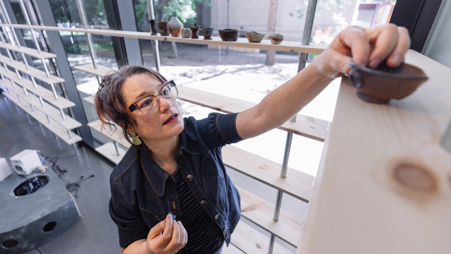 An artist carefully arranges small pottery pieces on a wooden shelf in a well-lit studio, showcasing their creative workspace.