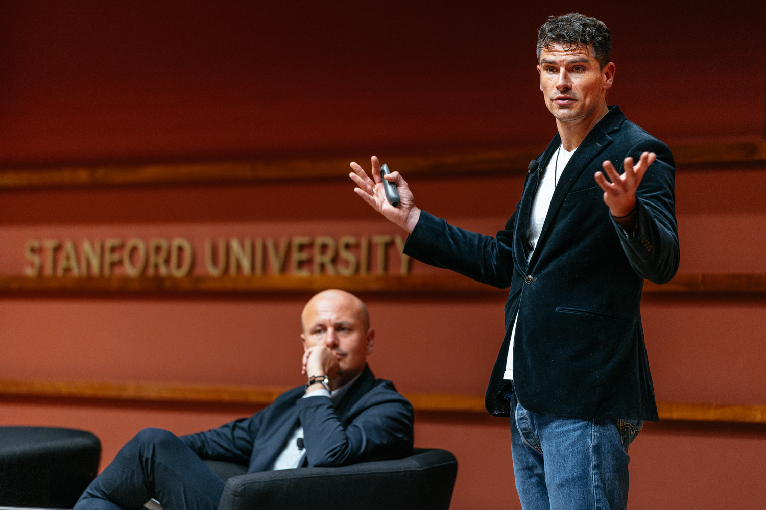 Luke Kemp in blazer gestures while speaking at Stanford University auditorium.