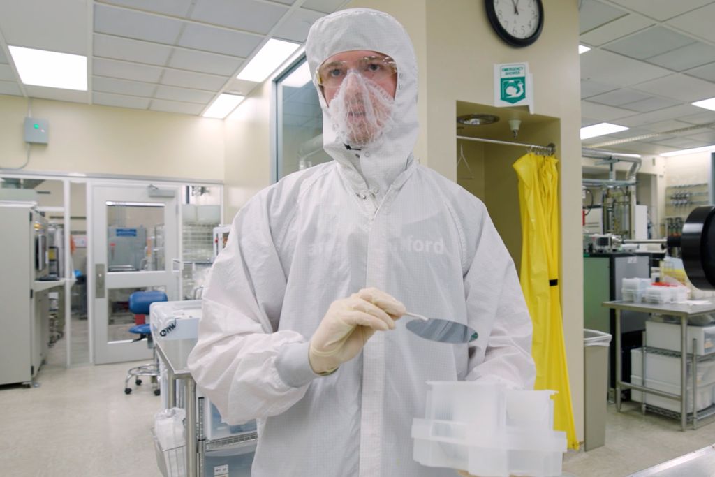Open video A lab worker in protective gear holds an item with prongs and a container while standing in a cleanroom environment. in a modal