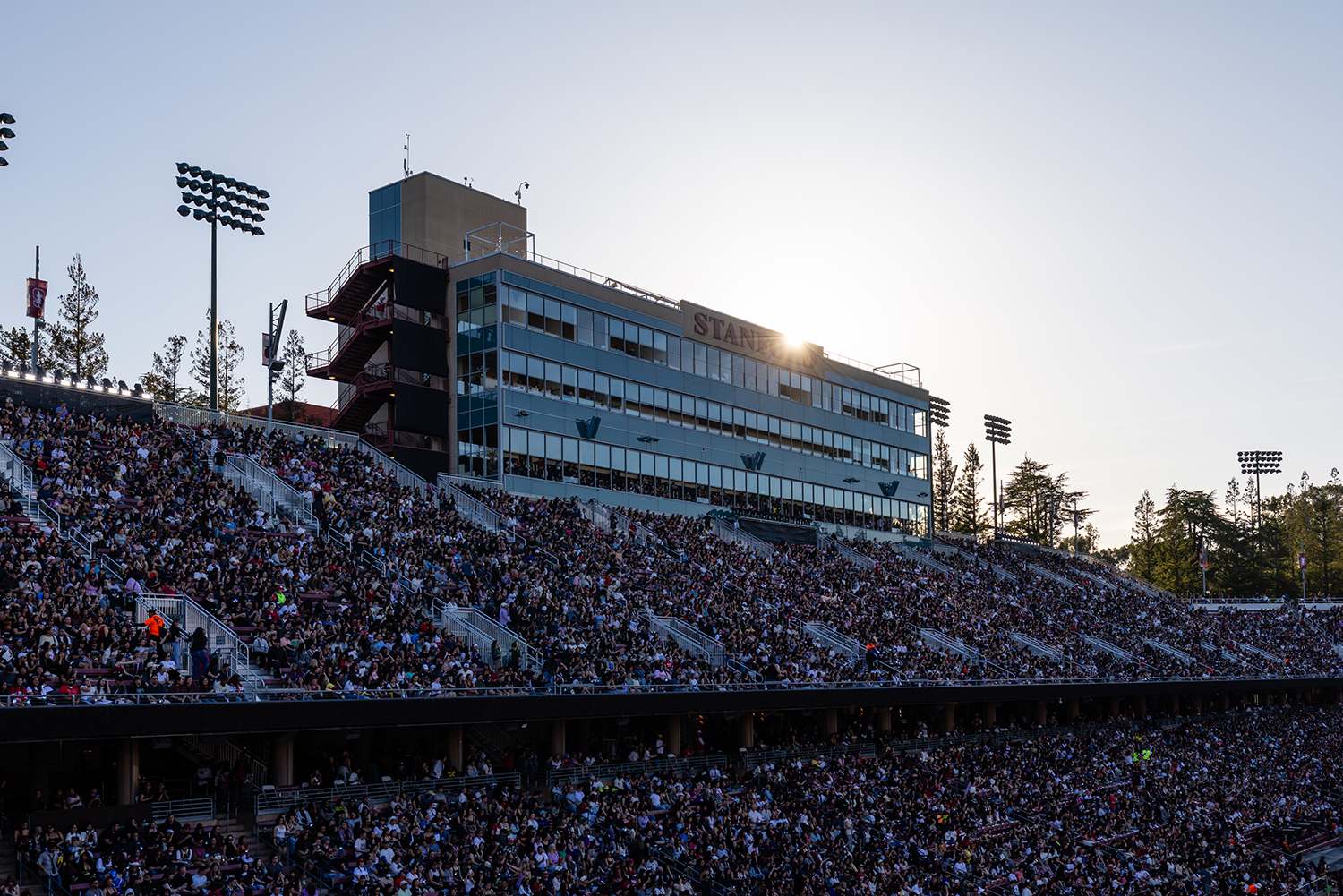 A large crowd fills the stands of a stadium, with the setting sun shining over the building in the background.