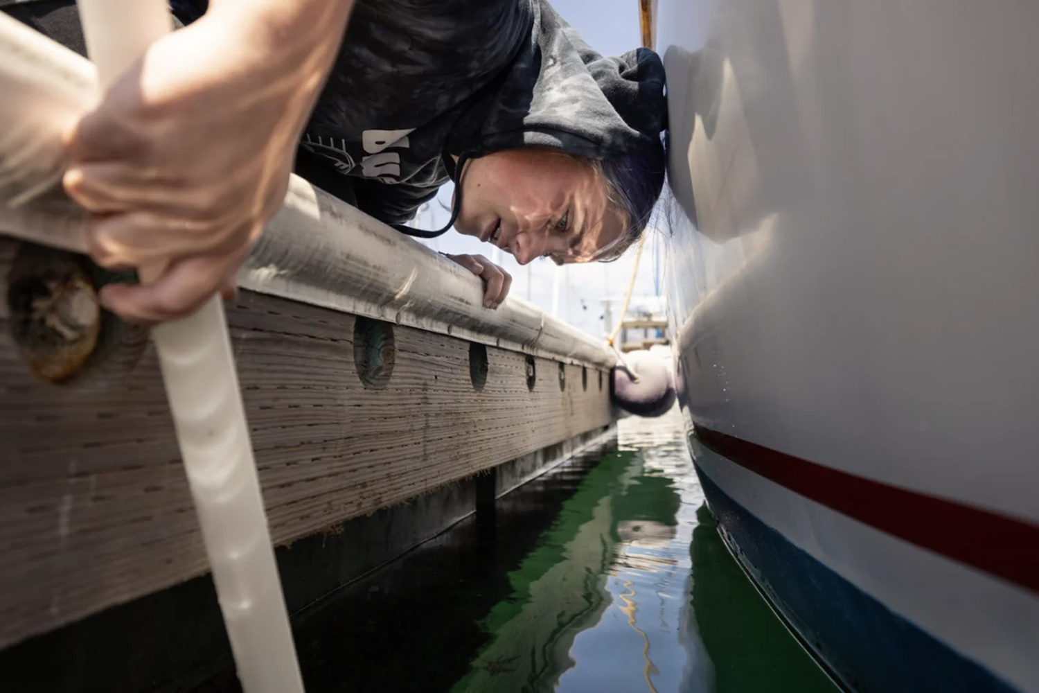 A person reaches over a dock to interact with the water alongside a boat, with reflections visible in the calm water.