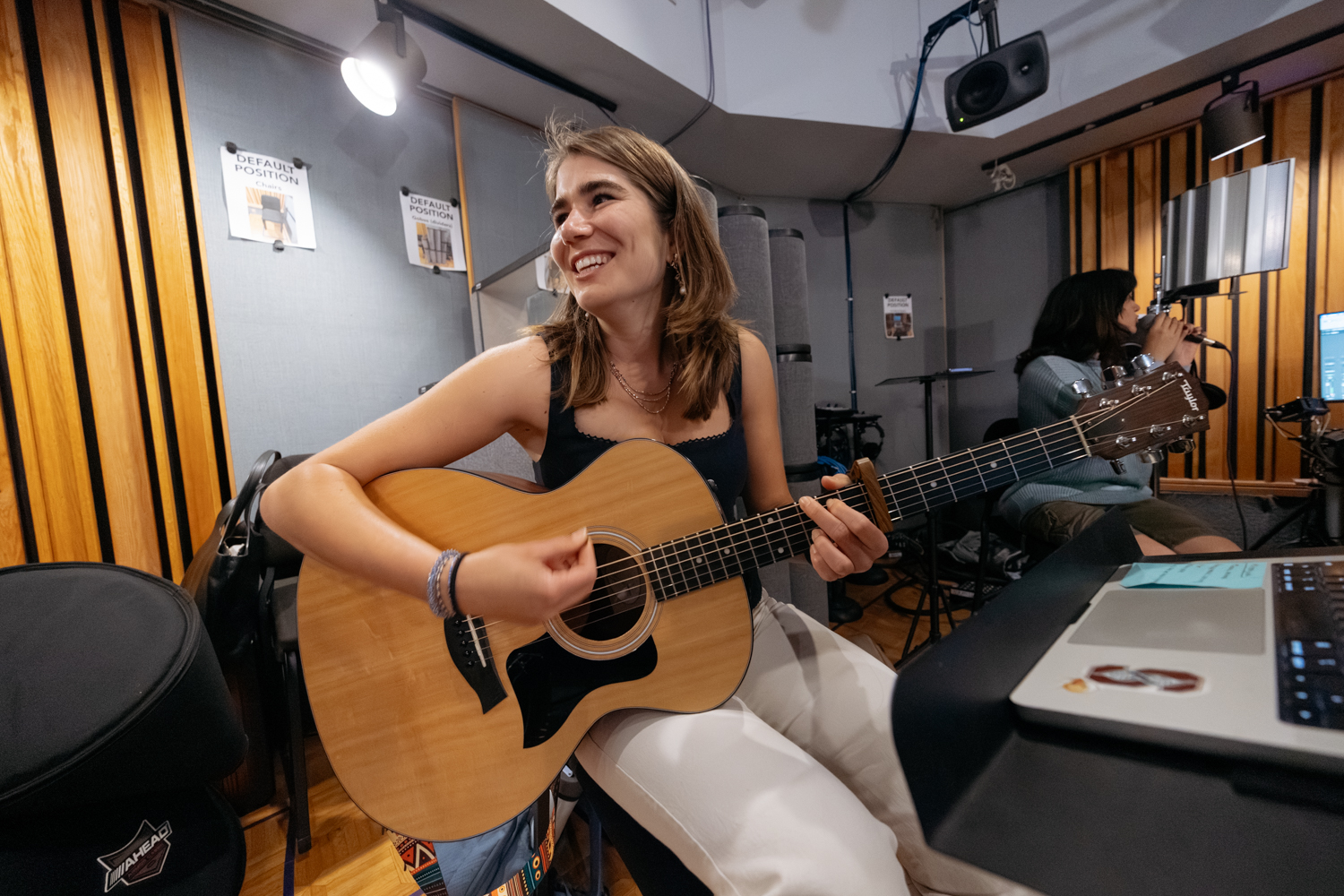 Hoffmann plays an acoustic guitar while seated in a studio, with another person singing nearby.