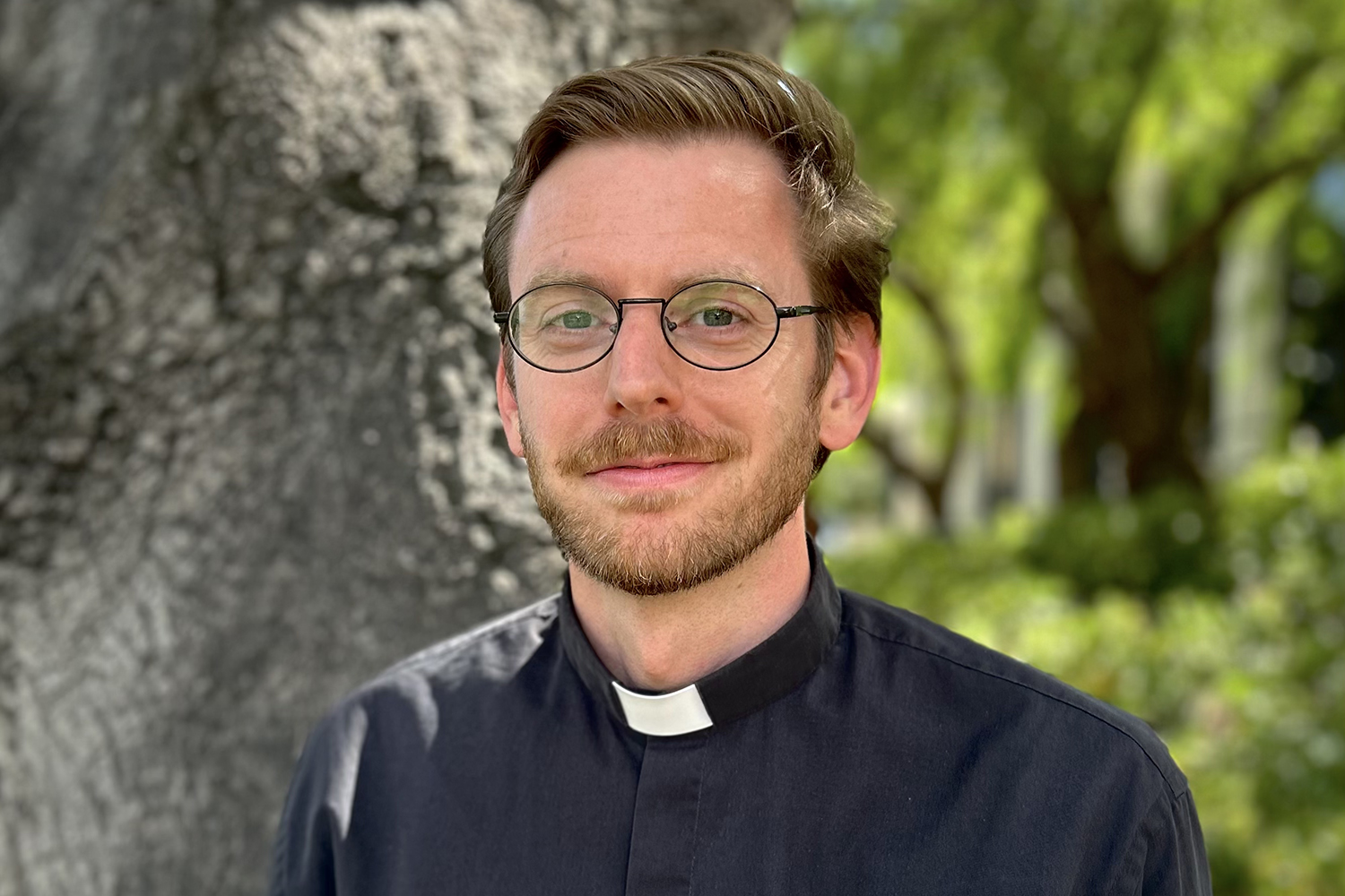 Thomas wearing glasses and clerical collar standing outdoors with tree and foliage in background.