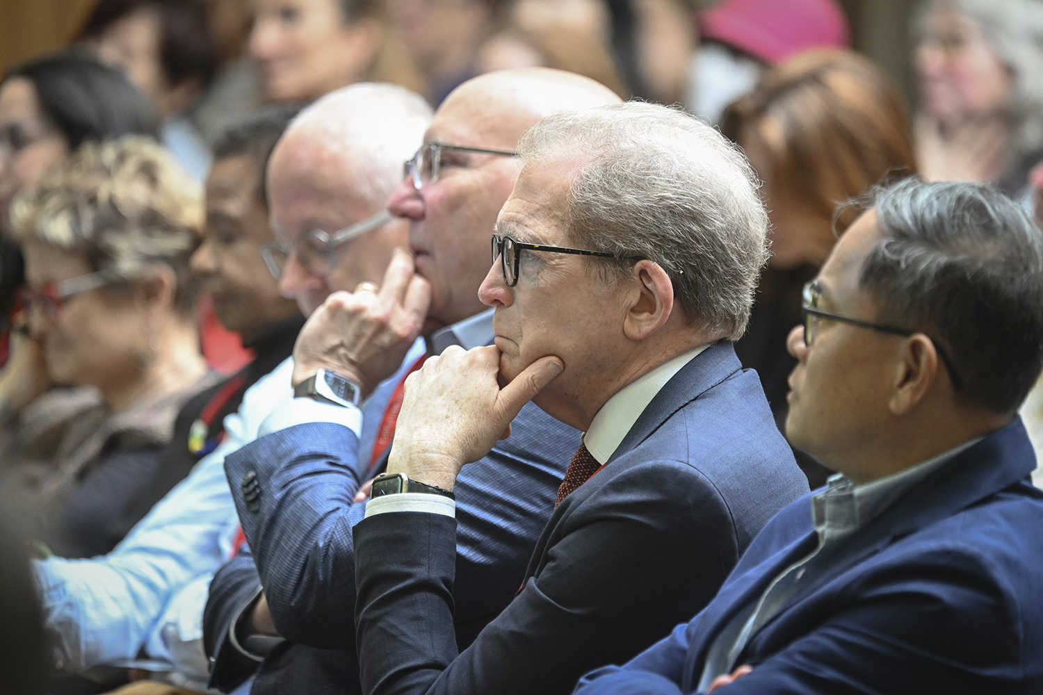 Dean Minor and a diverse group of people sits in the audience, attentively listening to a speaker at an event.