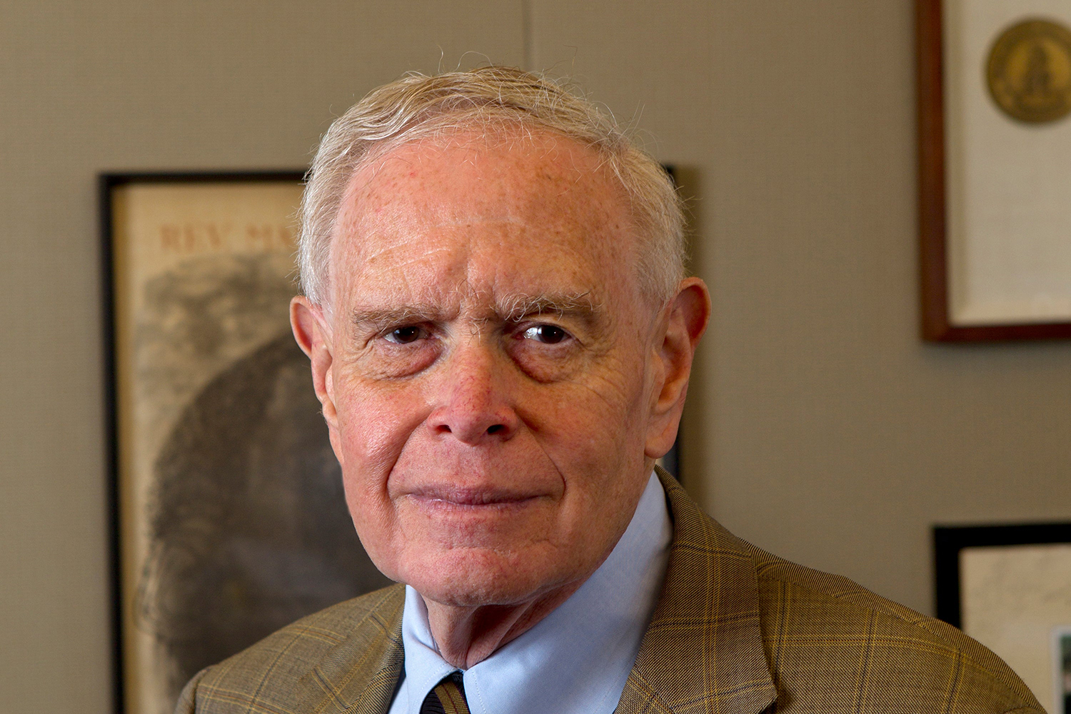Paul Brest in a checked blazer and blue shirt stands indoors against a backdrop of framed objects.
