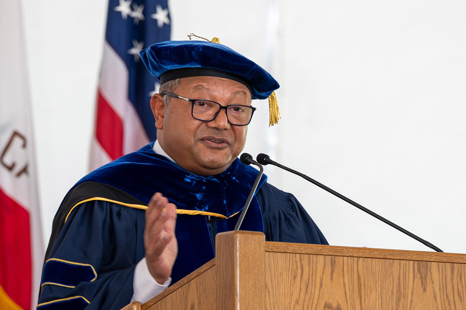School dean Arun Majumdar in academic regalia addresses the audience from a wooden lectern during a graduation ceremony.
