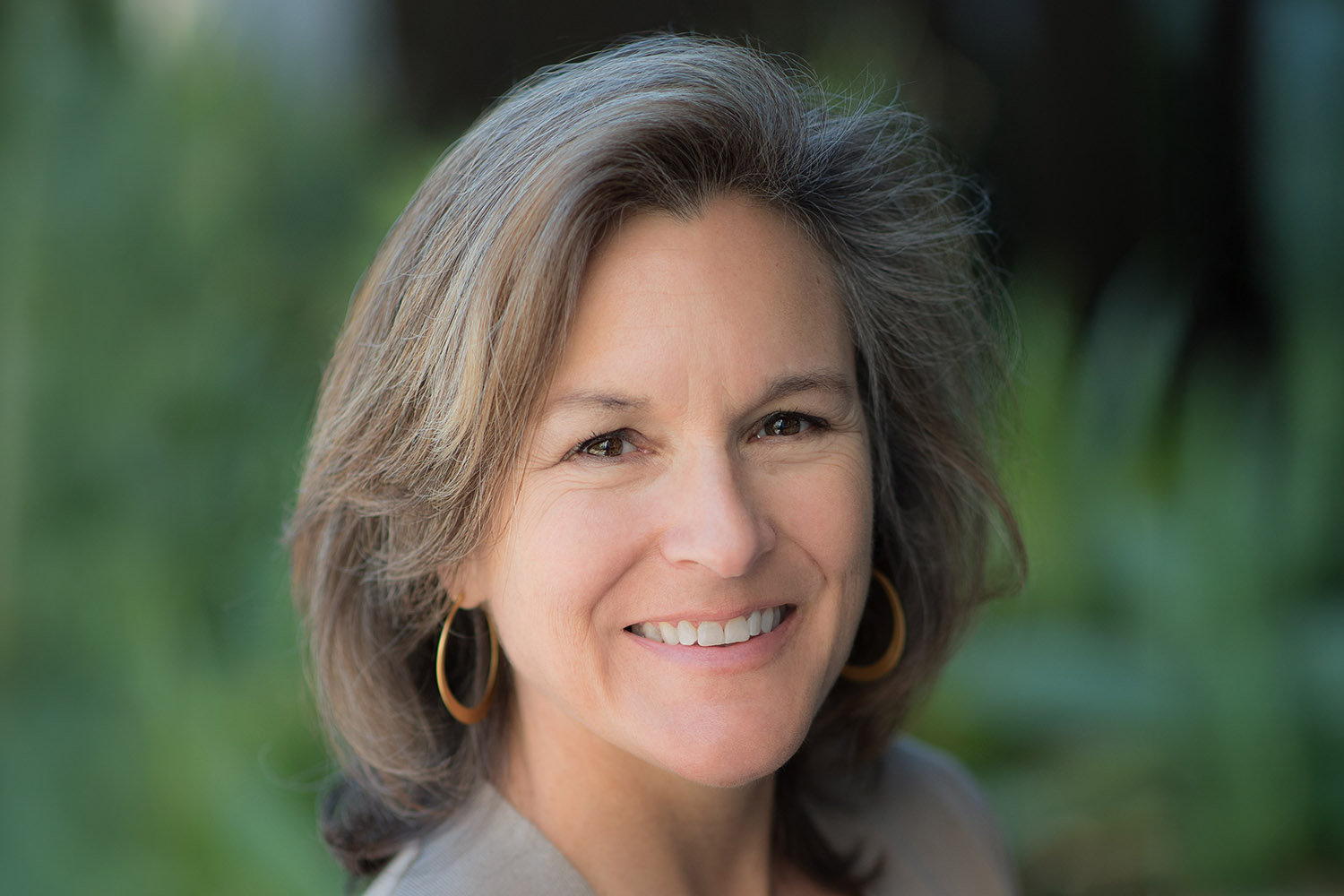 Headshot of Tracey Woodruff with shoulder-length hair and golden hoop earrings posed outdoors against a blurred green background.