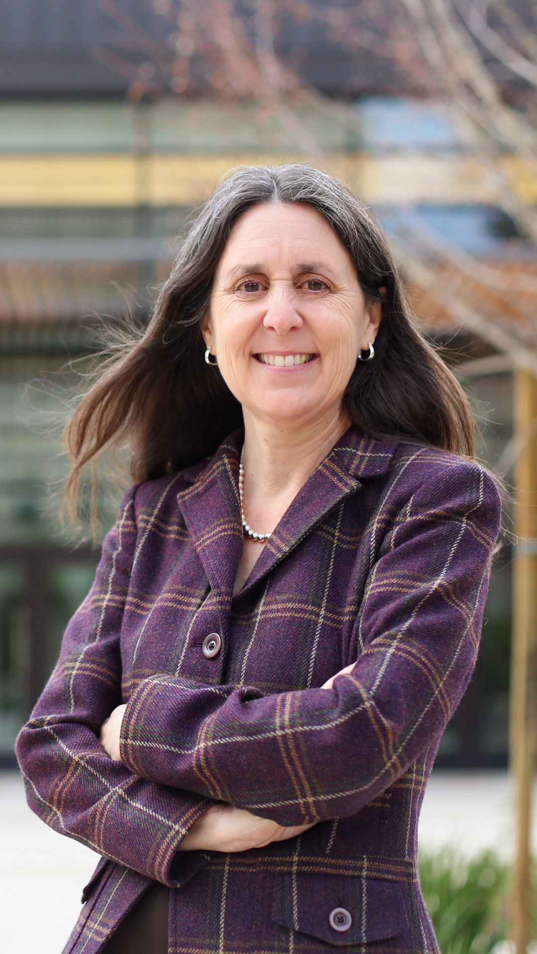 Jennifer Wolf with long hair smiles confidently while wearing a plaid purple blazer, standing outdoors with a blurred background.