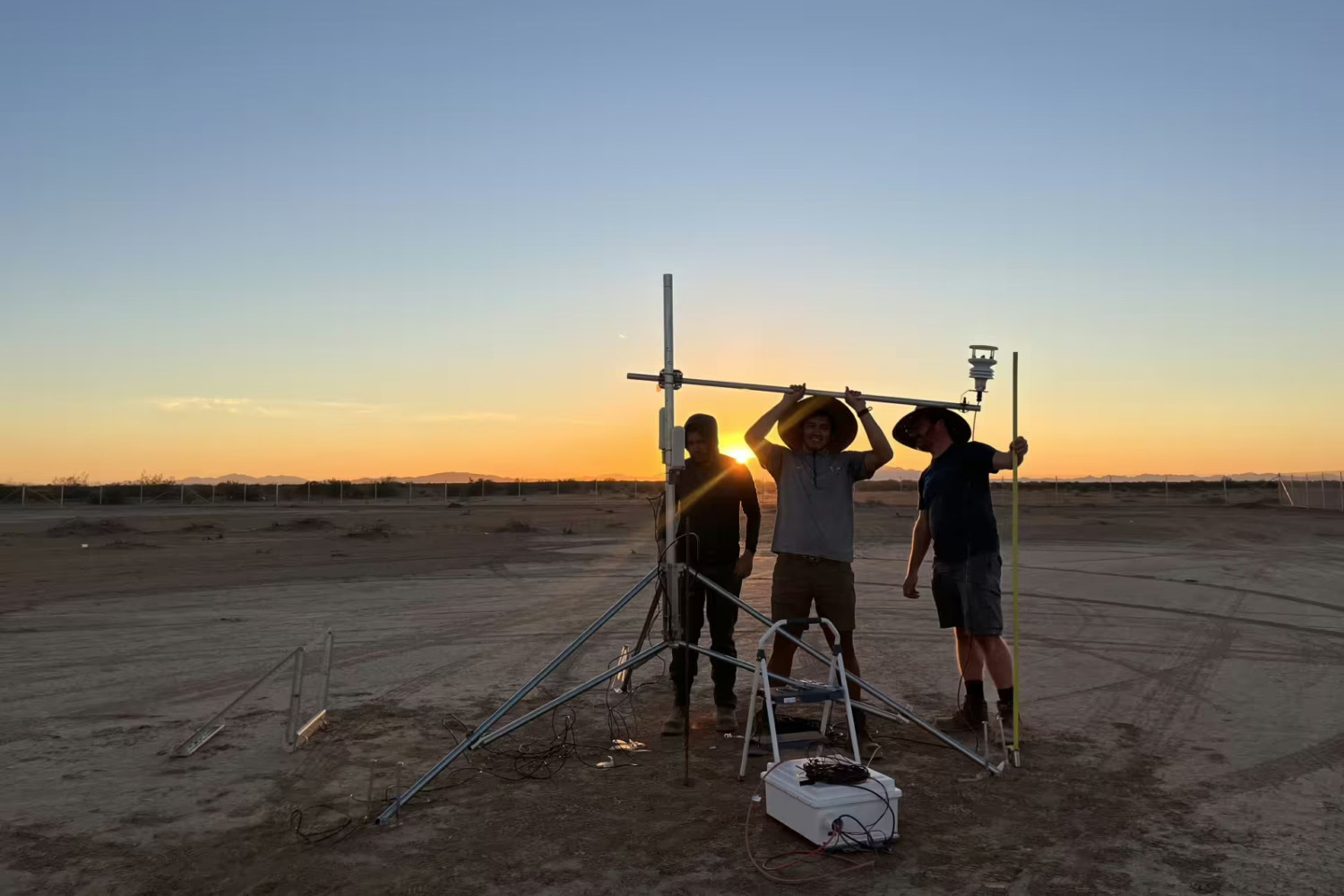 Researchers Adam Brandt, Josh Romo, and Taylor Adams set up a tripod-mounted weather sensor on a desert flat at sunset.