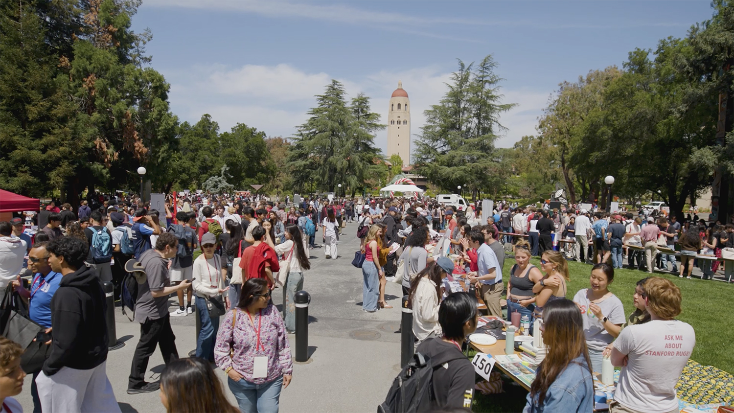 Large outdoor campus event with crowds gathered near tables and tents, Hoover Tower visible in the background.