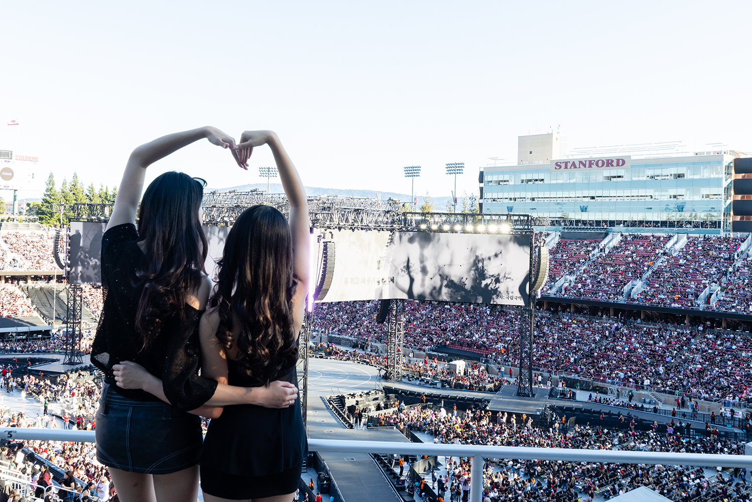 Two friends make a heart shape with their arms and hands while looking at a concert stage and a crowded stadium.