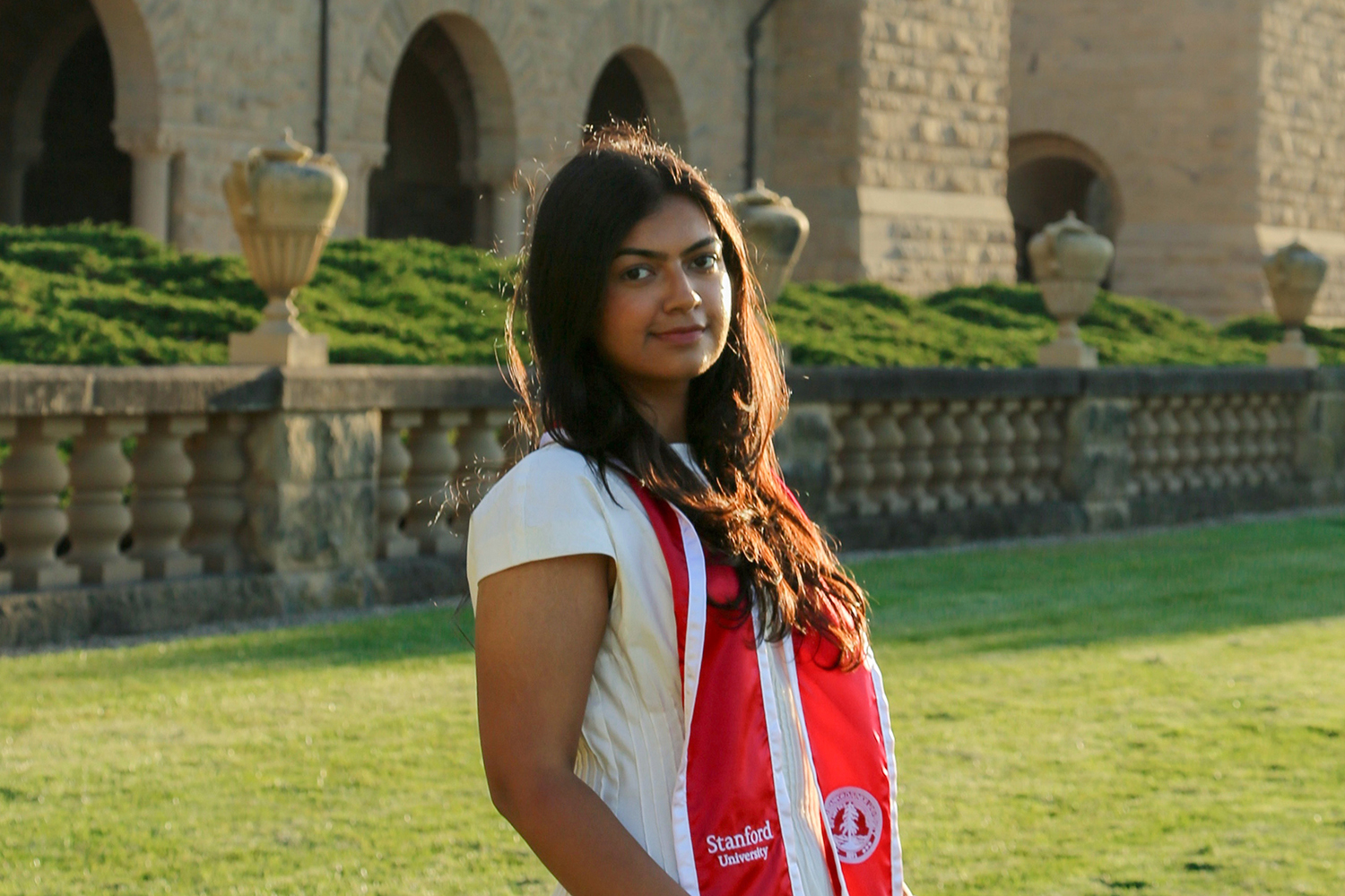 Lamya Butt Butt in a white dress and red graduation stole stands outdoors, with Main Quad and greenery in the background.