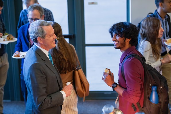 Randal Quarles and a student talk while people mingle in the background, holding plates of food.