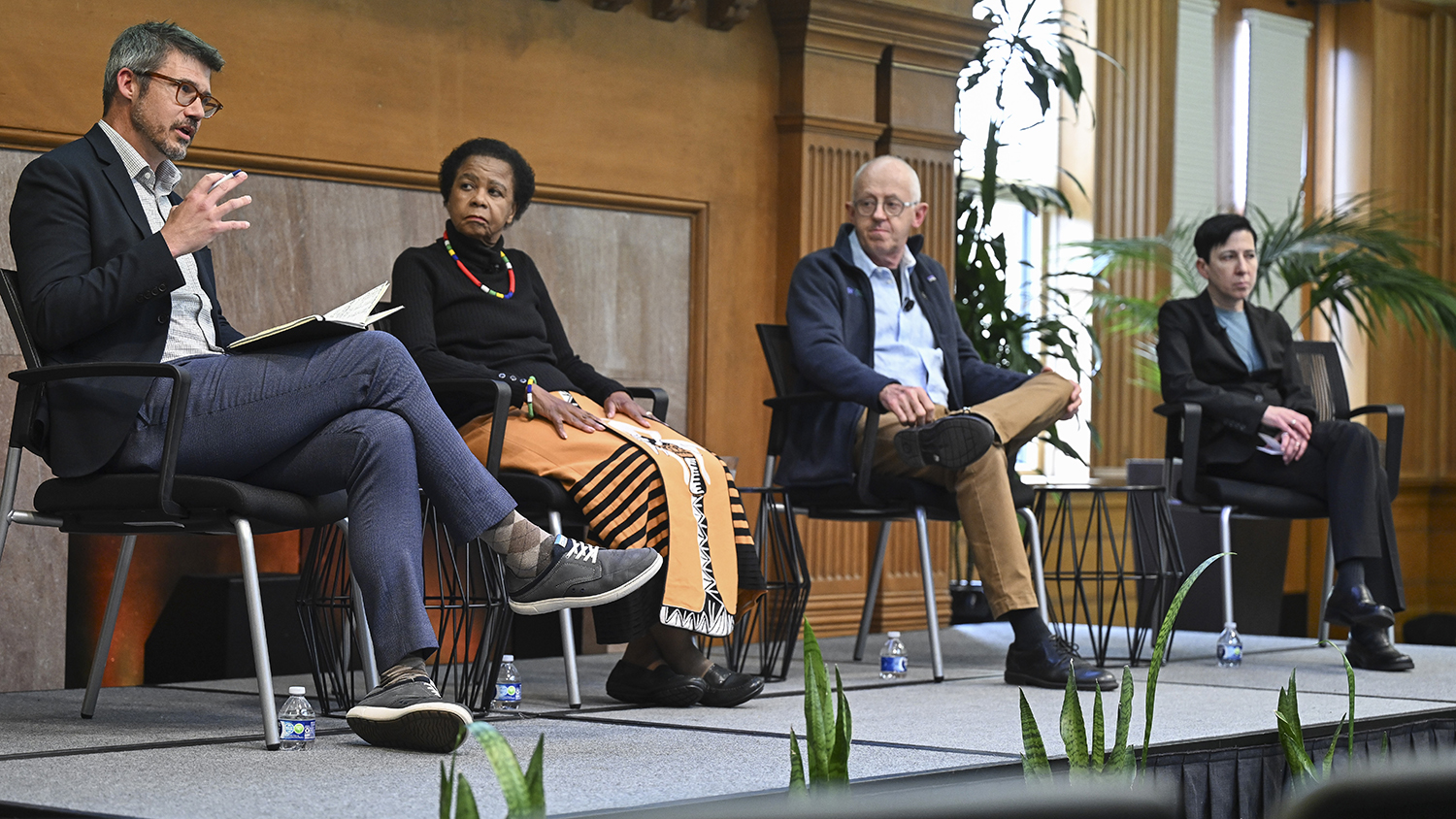 Four panelists sit on stage discussing a topic, with one actively engaging the audience while the others listen attentively.