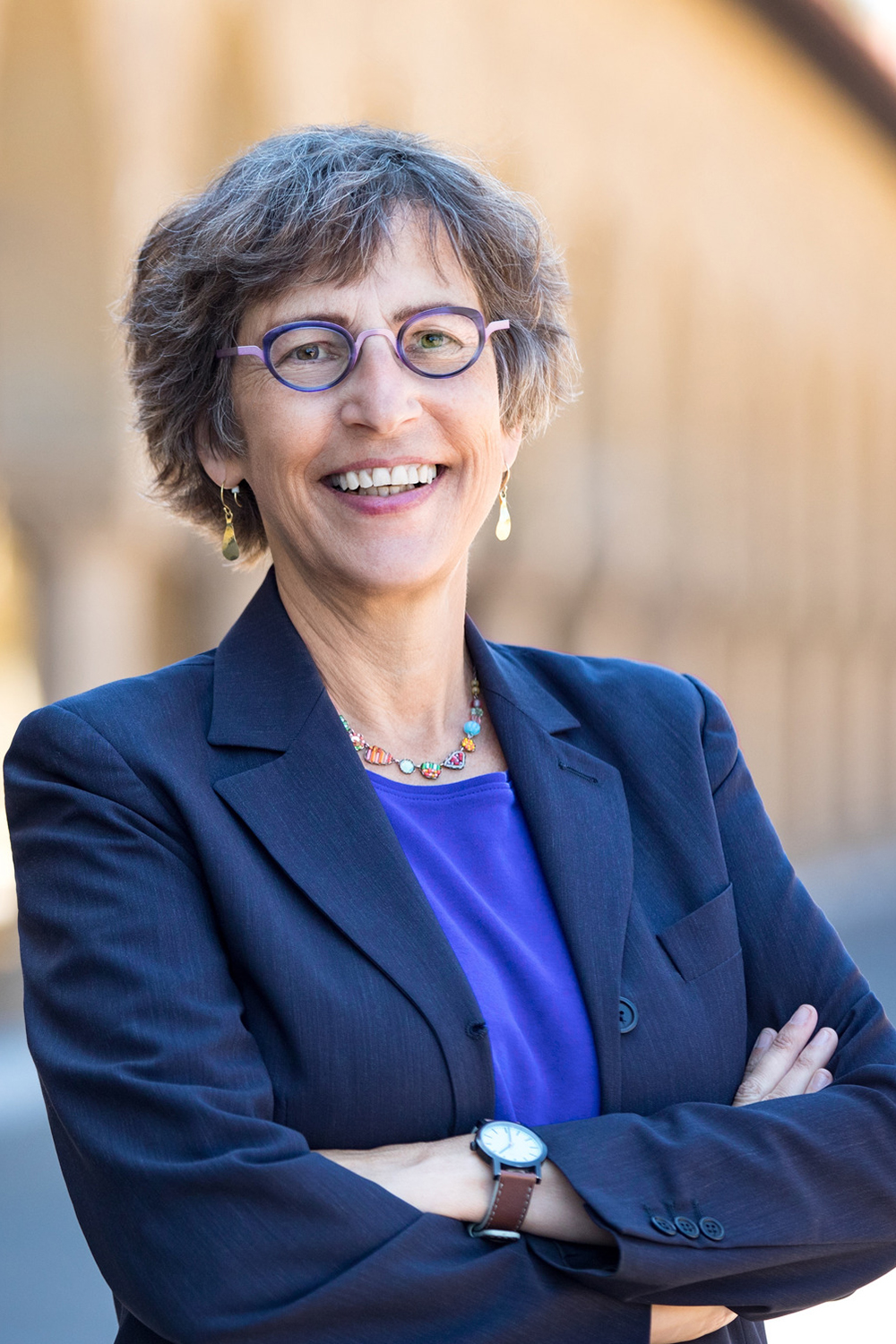 Debra Satz wearing a dark blazer, a colorful necklace, and purple glasses poses confidently outdoors with arms crossed.