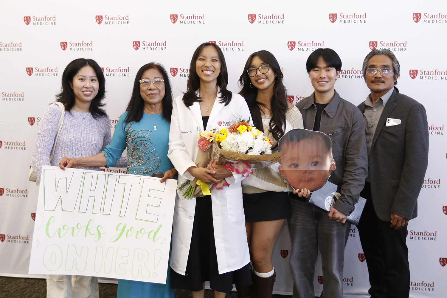 A medical student holds flowers while posing with family members holding a handmade sign and photo.