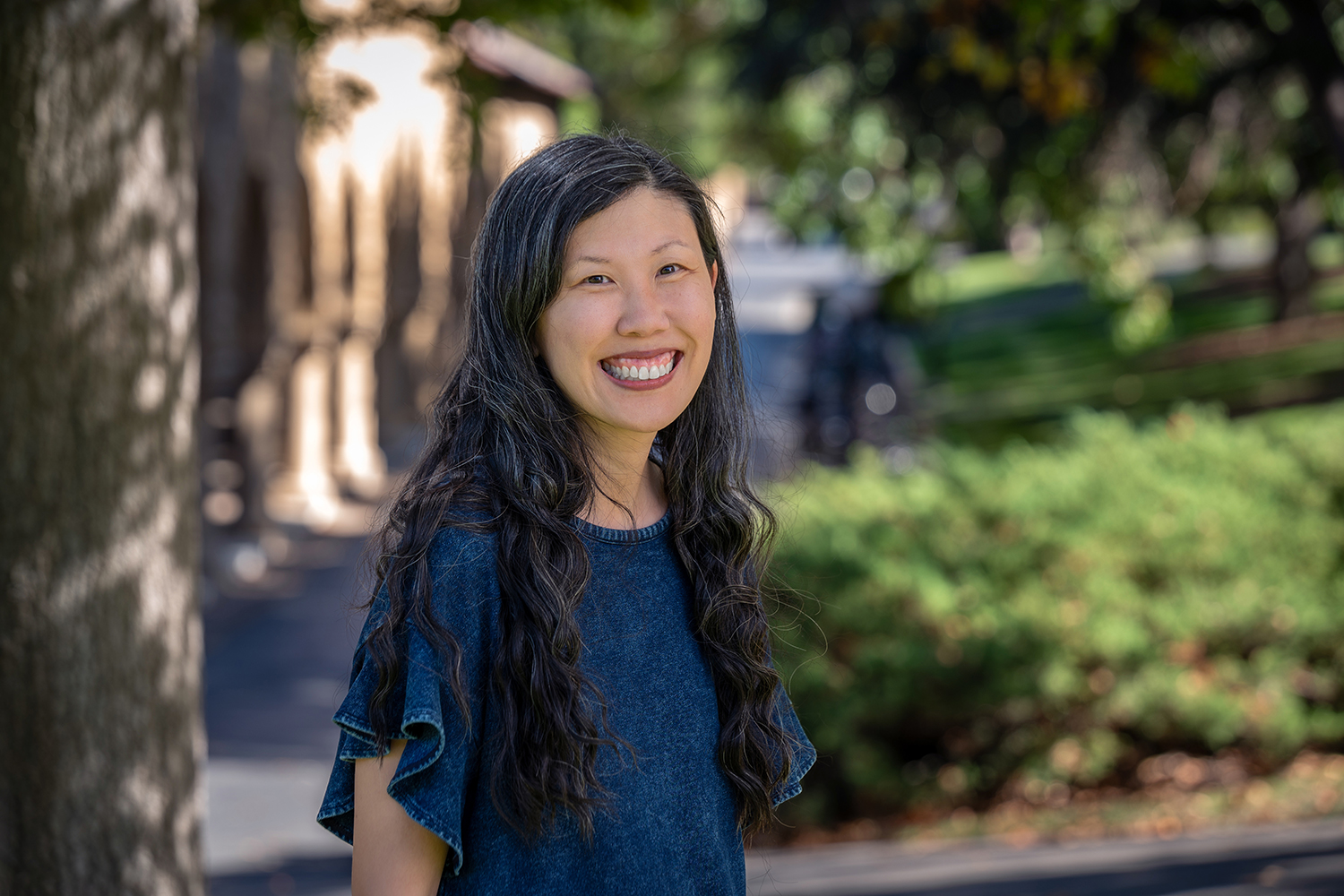 Wendy Mao stands outdoors and poses for a professional headshot in front of a blurred arcade and greenery.