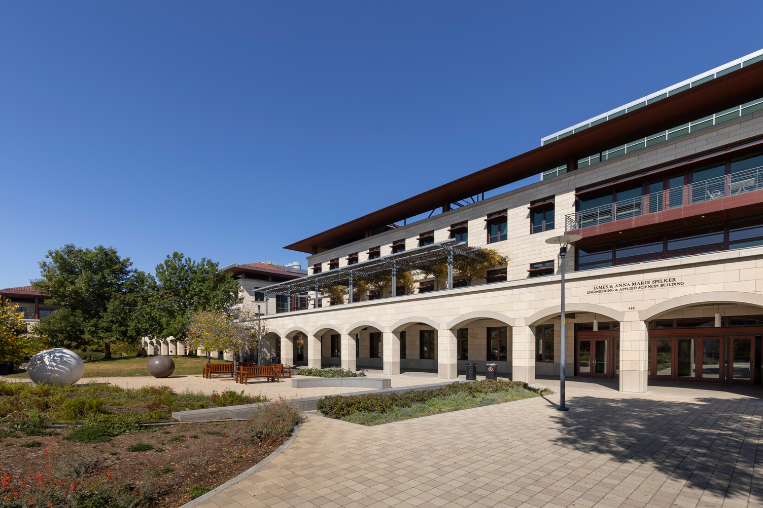 Spilker Engineering and Applied Sciences Building with a ground-floor arcade and red-trimmed upper floors, facing a paved courtyard with benches and stone sculptures.