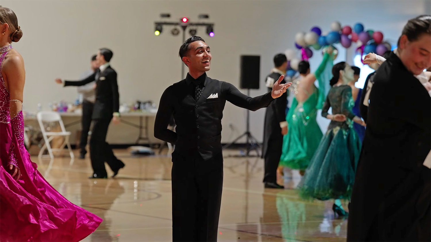 Dancers in formal attire perform in a ballroom, with colorful dresses and a festive atmosphere featuring decorations in the background.