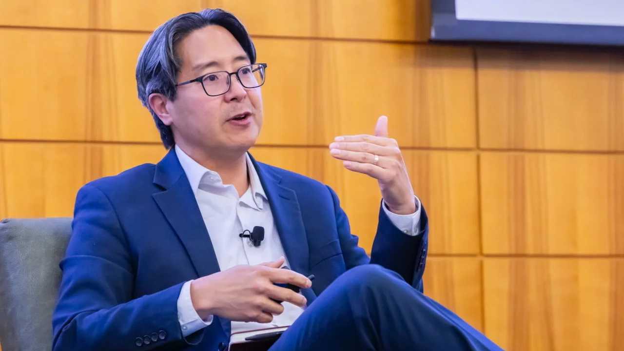 Michael Hsu in a suit speaks while seated in a chair, gesturing with one hand during a panel discussion.