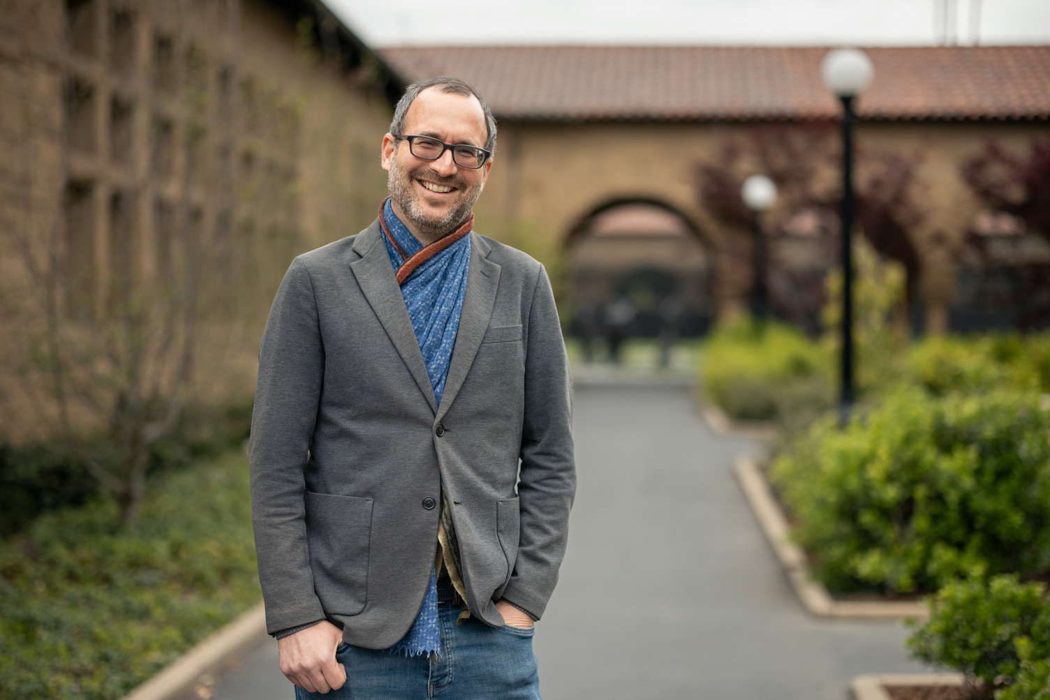 Dan Edelstein in gray blazer and blue scarf smiling on a campus walkway with historic buildings in the background.