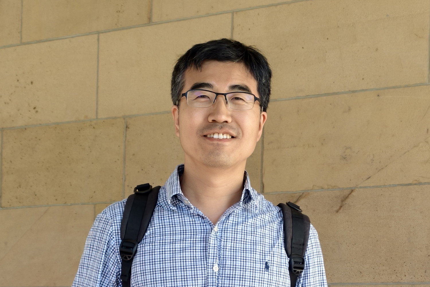 Xiao-Liang Qi wearing a backpack and posing for a photo in front of a sandstone block wall.