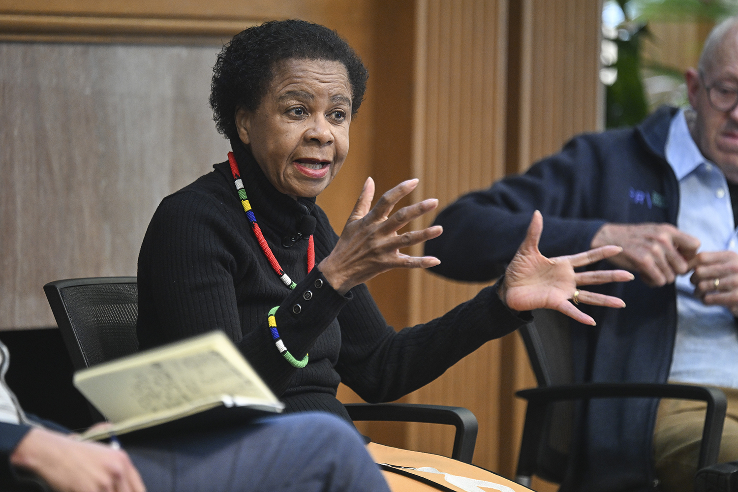 Mamphela Ramphele gestures while speaking at a panel discussion, with two seated individuals visibly engaged in the conversation.