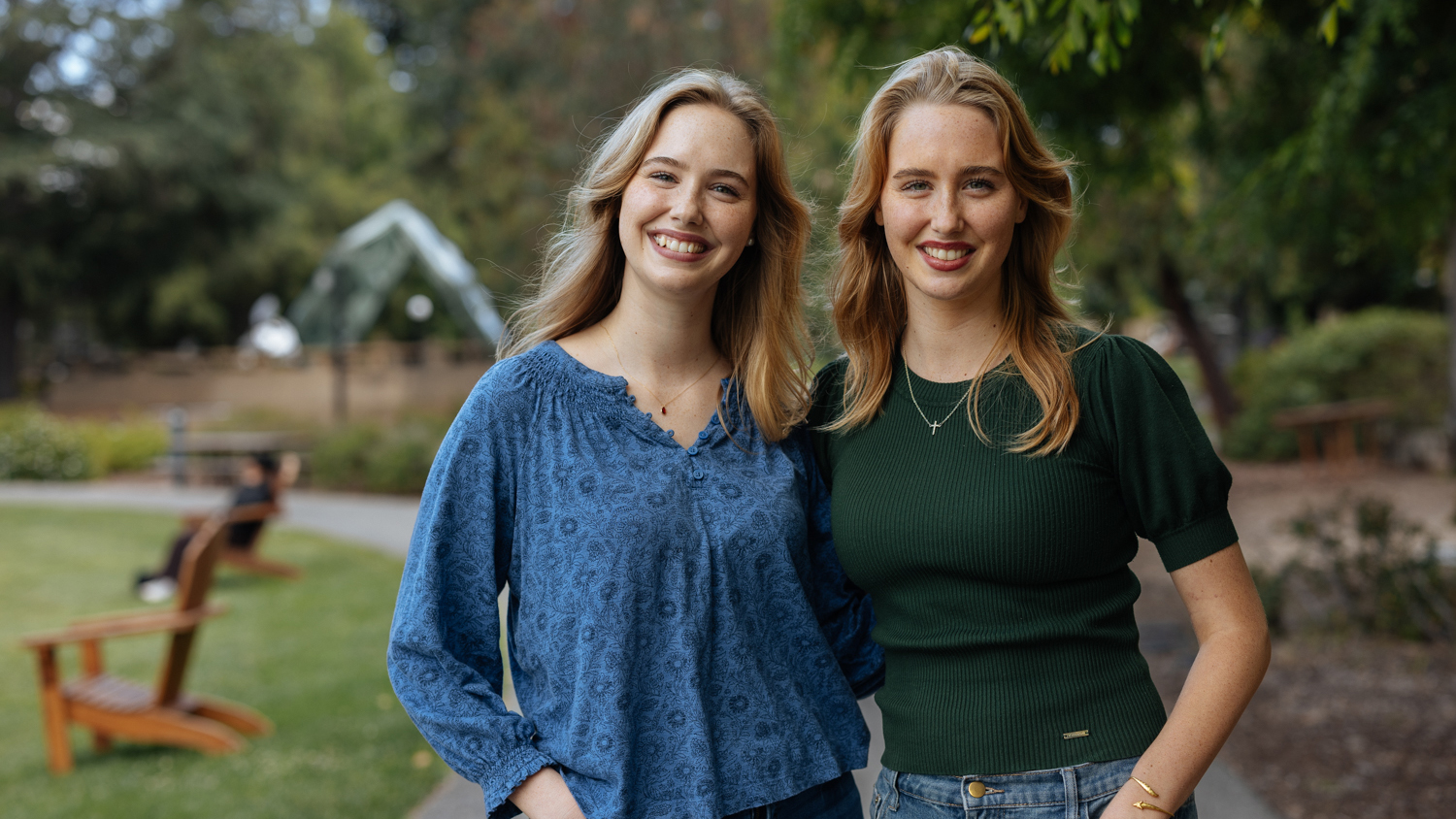 Twin young women with long blond hair stand outside smiling on a sunny day.