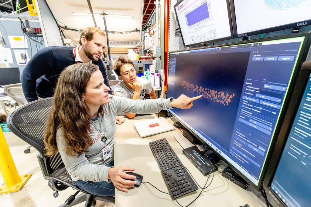 A group of people examines data on multiple computer screens in a laboratory setting.