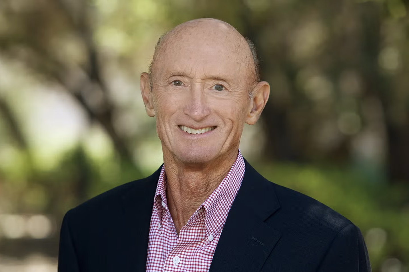 Holloway smiling, in a blazer and checked shirt, is posing outdoors, with blurred trees in the background.