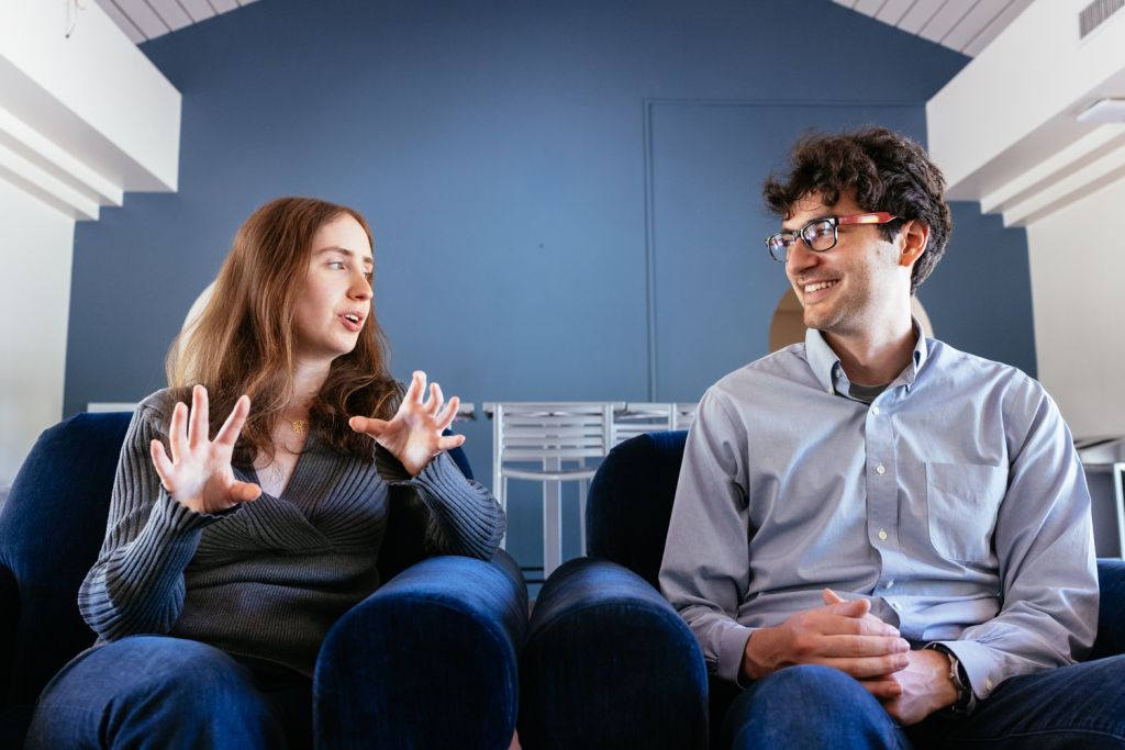 Julia Proshan and Daniel David Richman sit in a casual setting, with Julia gesturing as they discuss a topic, conveying engagement and communication.