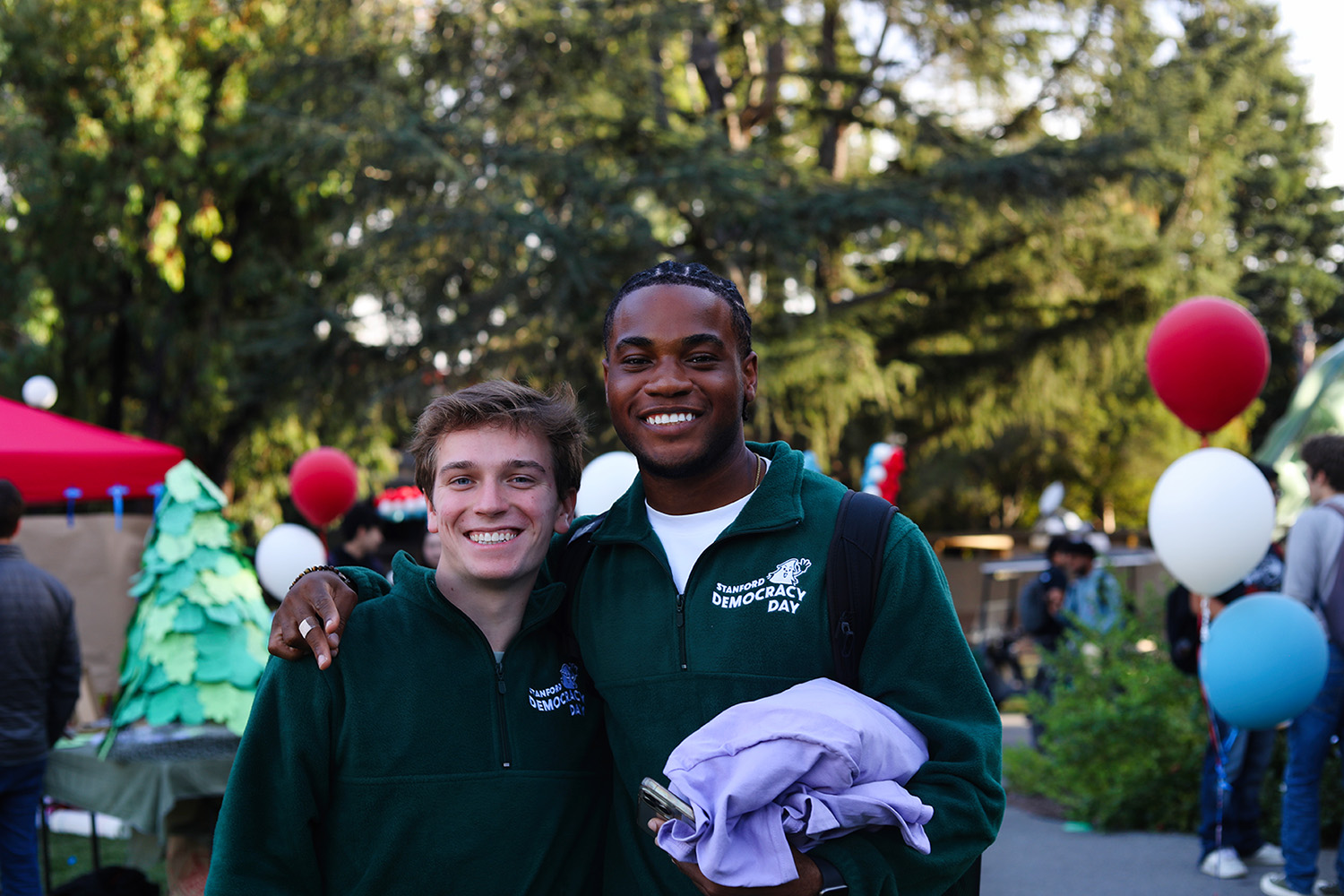 Jadon Urogdy and Uchenna Akanno in matching green fleeces with arms around each other at an outdoor Democracy Day event, balloons behind them.