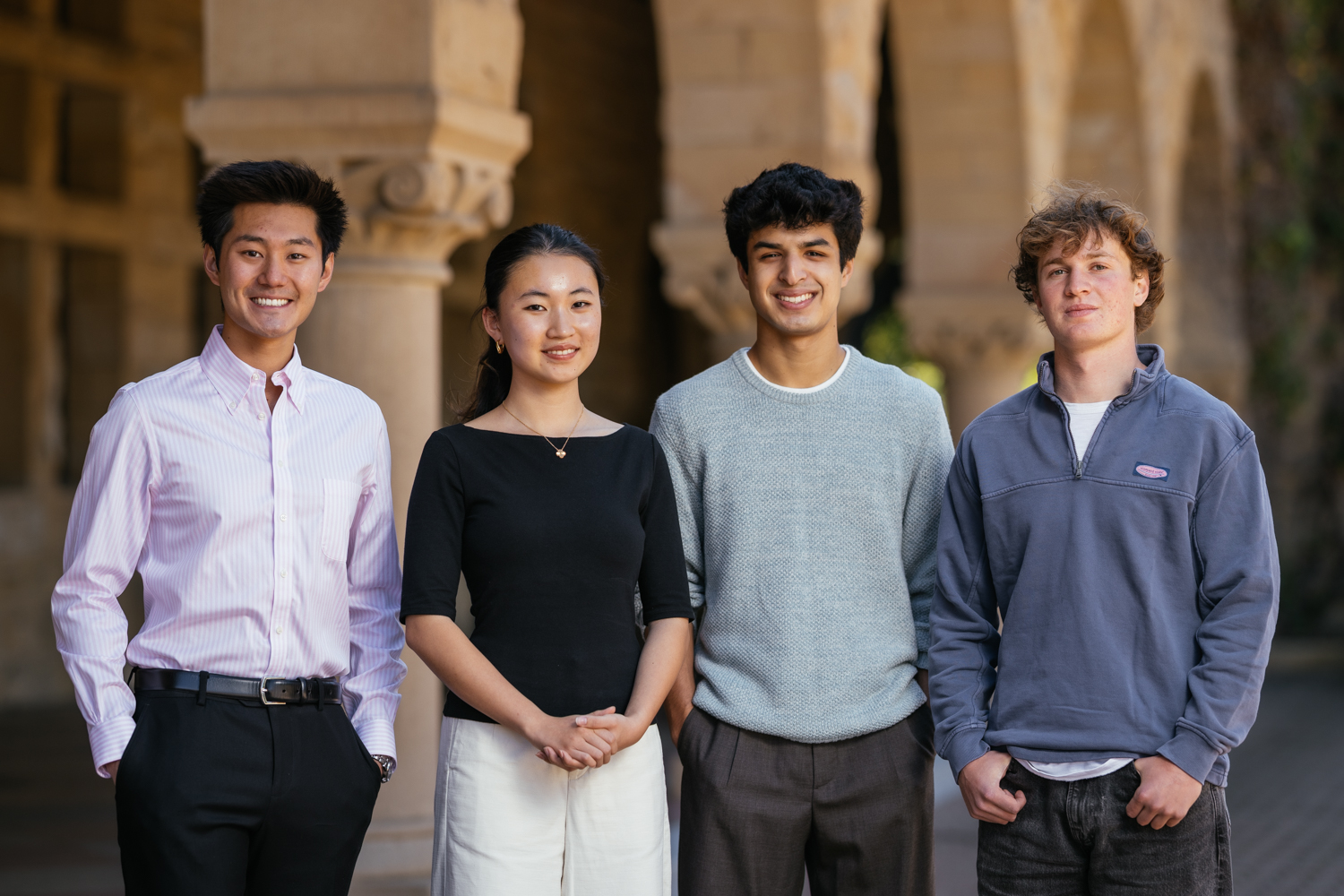 Four students stand together outdoors, smiling and wearing casual clothing against Quad arcade backdrop.