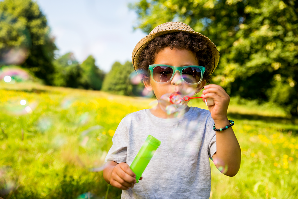 Male child wearing a hat and sunglasses blowing bubbles in a park setting.