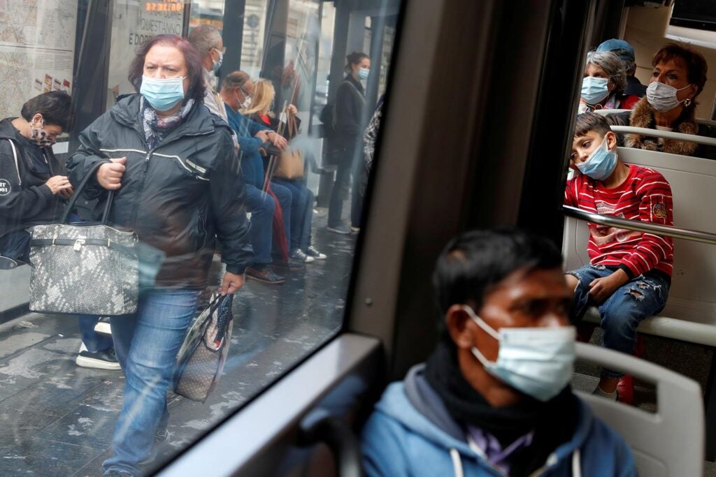 Masked passengers sitting in a public bus, and a person seen through the bus window with shopping bags, also wearing a mask.