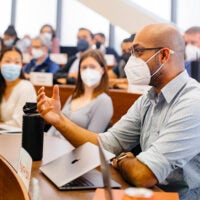 GSB students in a classroom wearing masks, watching intently as a male student holds his hand forward while speaking to the class.