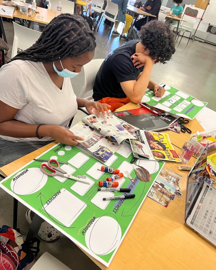 2 students sitting at a table with magazines and poster boards, creating collages.