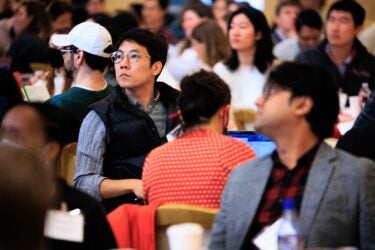 Image shows crowded meeting room and focuses on one conference participant looking up deep in thought while listening to a speaker.