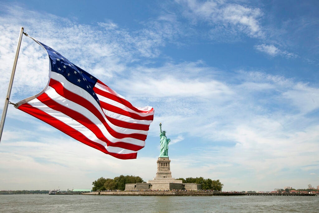 The statue of liberty in the center of the frame, with an American flag waving in the foreground.
