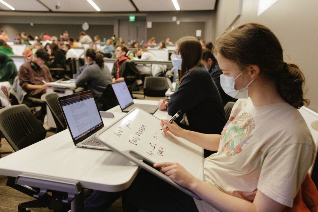 Student seated at a desk, handwriting formulas on a handheld dry erase board.
