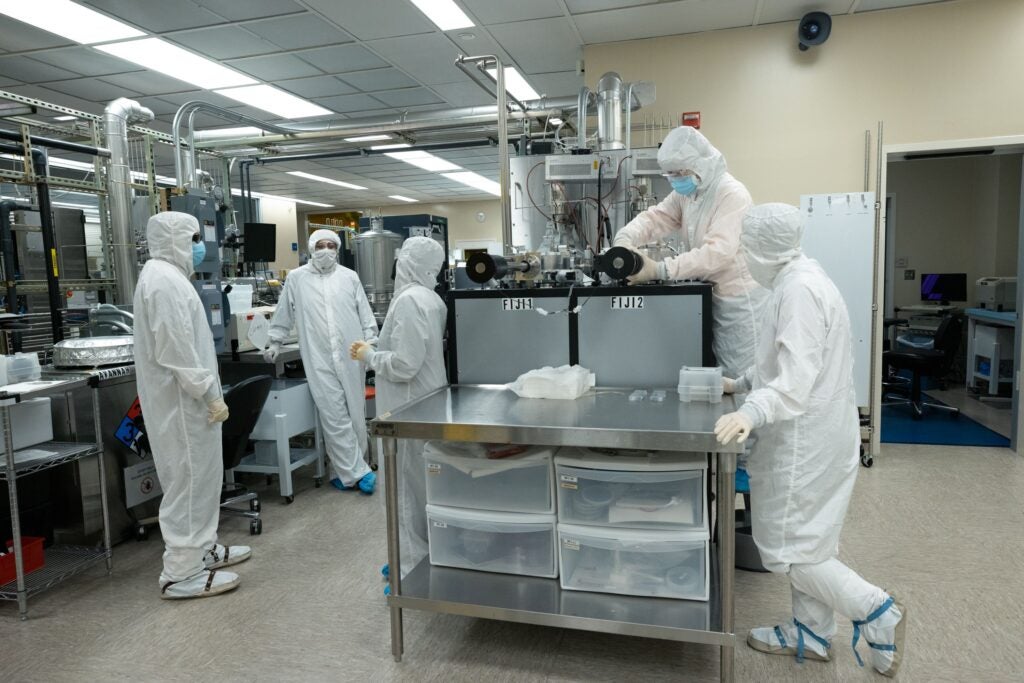 Five people in clean room suits. Three are talking while two work on equipment.